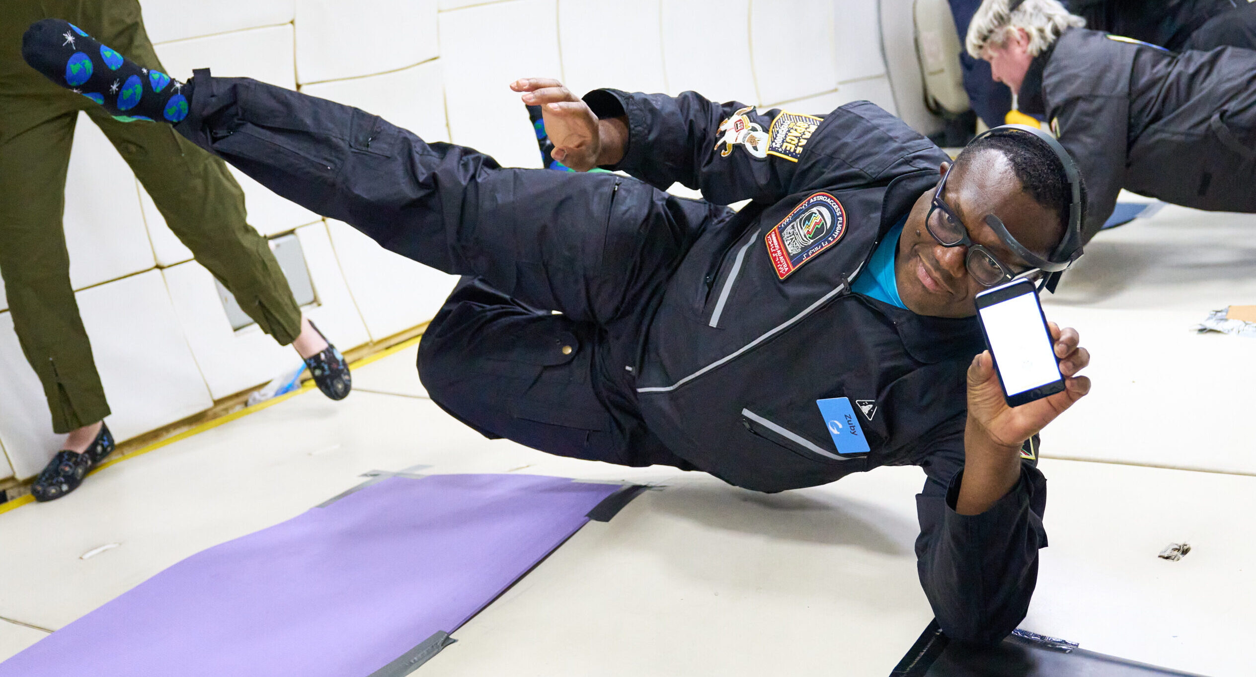 Zuby, a low vision ambassador, holds up his phone towards the camera as he floats in zero gravity, displaying a test for his heads-up display technology prototype.