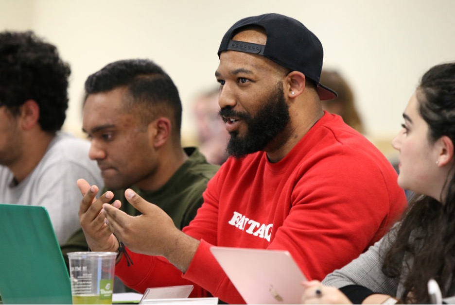AJ is black male wearing a backwards black hat and red sweater. He is sitting down next to other folks and speaking with expressive hand gestures. 