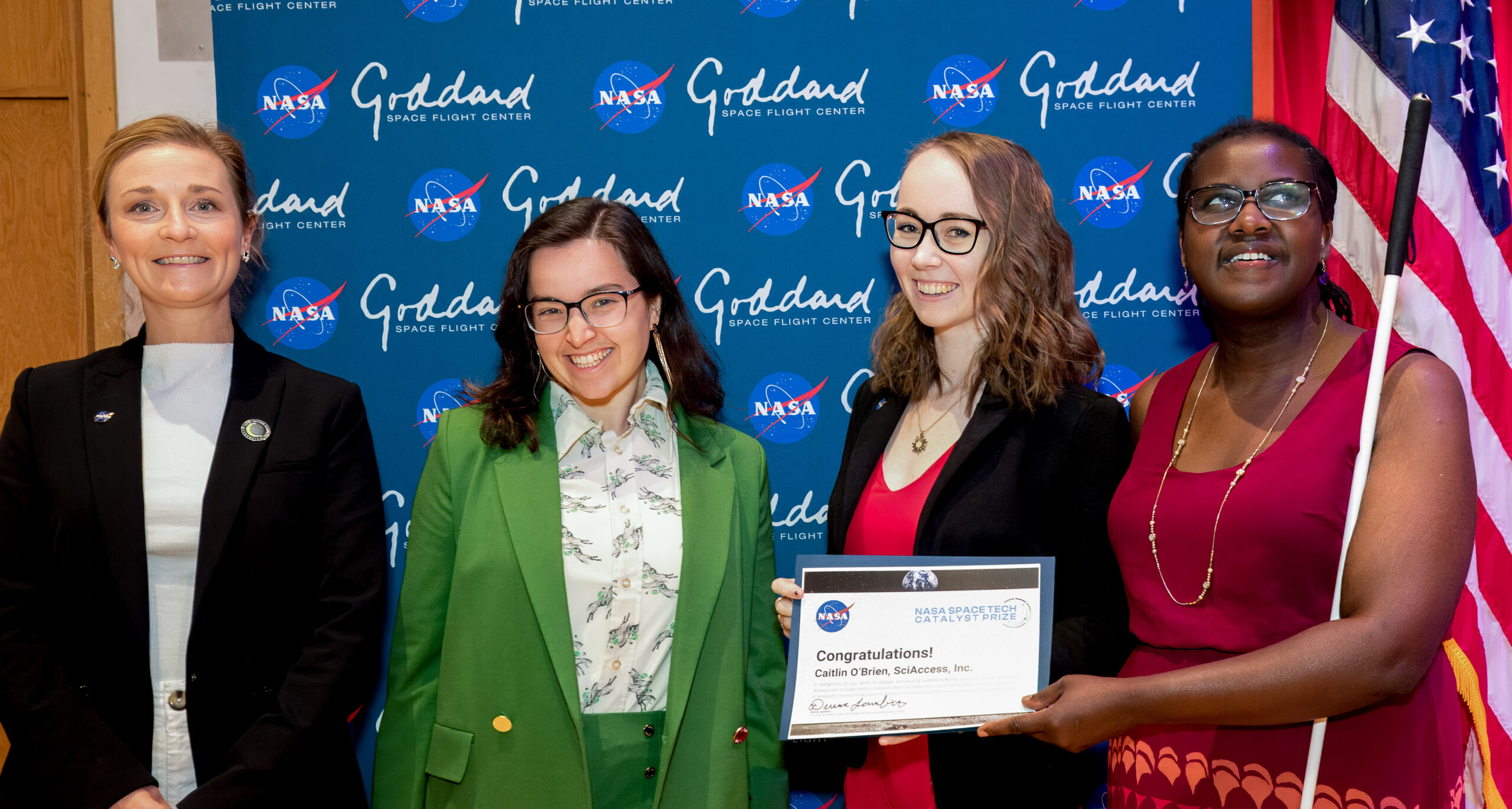 An image of four people smiling in front of a NASA Goddard backdrop. From left to right: Jenn Gustetic, Anna Voelker, Caitlin O'Brien, and Denna Lambert. Caitlin holds an award certificate and Deena is holding a white cane.