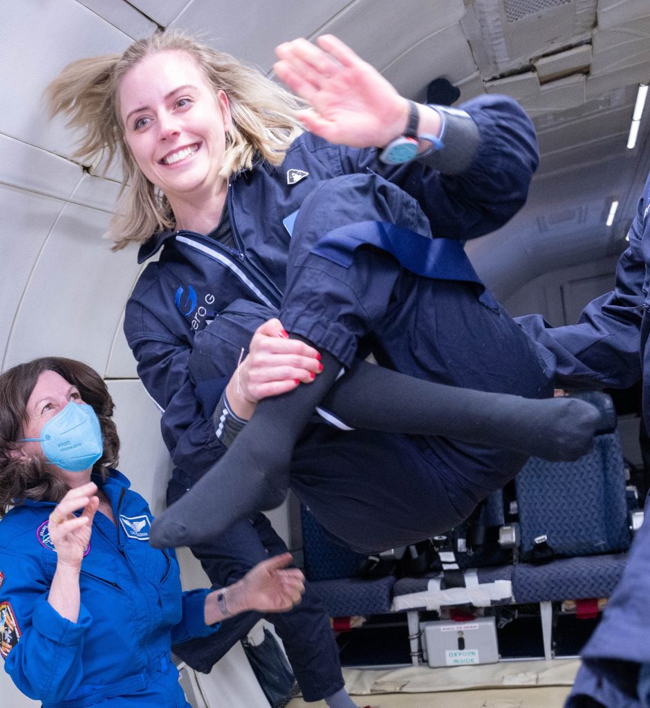 A smiling woman in a blue flight suit floats weightlessly inside an aircraft during a zero-gravity flight, waving while another crew member in a flight suit and mask steadies them from below.