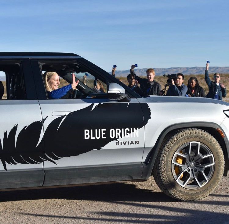 A woman riding a Blue Origin–branded Rivian SUV smiles and waves out the window while a group of people stand nearby cheering and photographing the moment against a clear blue sky and desert landscape.